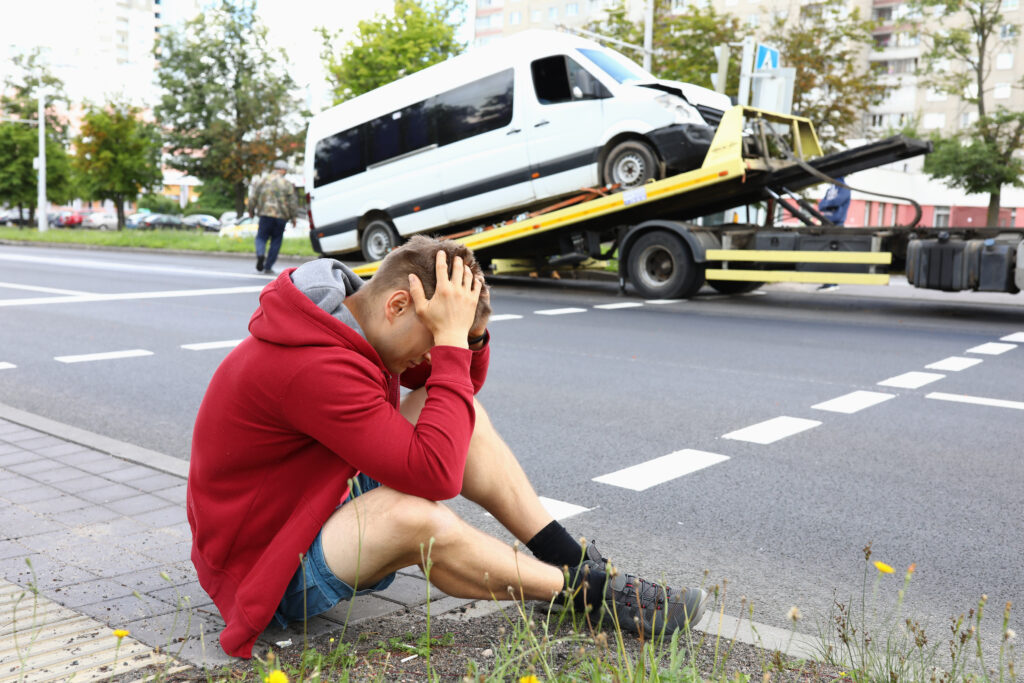Minivan driver sits on side of road and holds his head during evacuation of his vehicle after a car accident. Tow truck service business concept.